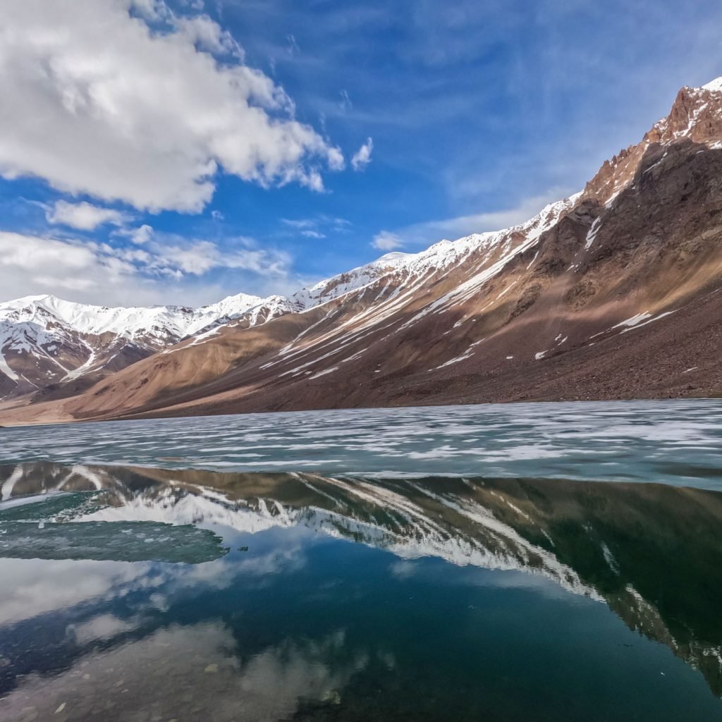Pristine Chandratal Lake with snow-capped Himalayan peaks and clear blue sky reflected in calm waters.