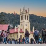A bustling scene in Shimla with people gathered around a prominent church featuring a clock tower, set against a backdrop of dense pine trees and a clear twilight sky.