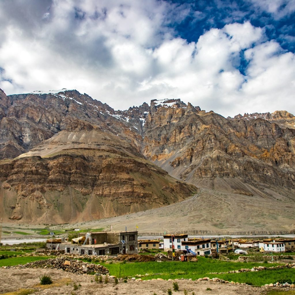 View of Kaza town nestled in the Himalayas with traditional buildings surrounded by green fields, a winding river, and towering stratified mountains under a partly cloudy sky.
