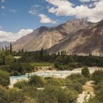 A serene view of Turtuk with a meandering turquoise river amidst lush greenery, flanked by towering brown mountains under a clear sky.