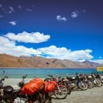 A scenic view of Tso Moriri lake with a row of parked motorcycles in the foreground, surrounded by barren hills under a bright blue sky with fluffy clouds.