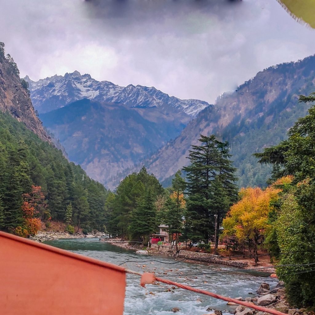 A serene view of the Parvati River flowing through a valley flanked by dense deodar trees and rugged mountains in the background under a partly cloudy sky.