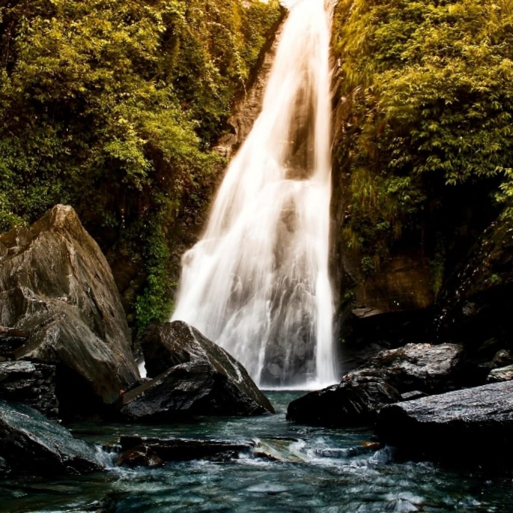 A majestic view of Bhagsu Waterfall with water cascading down a steep cliff surrounded by lush greenery and large rocks at the base.