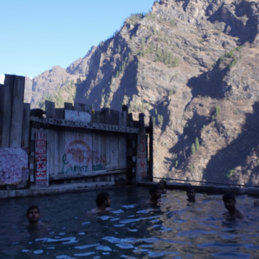 A natural hot water spring in Kheerganga trek with people bathing, surrounded by wooden fences and mountains under a clear sky.