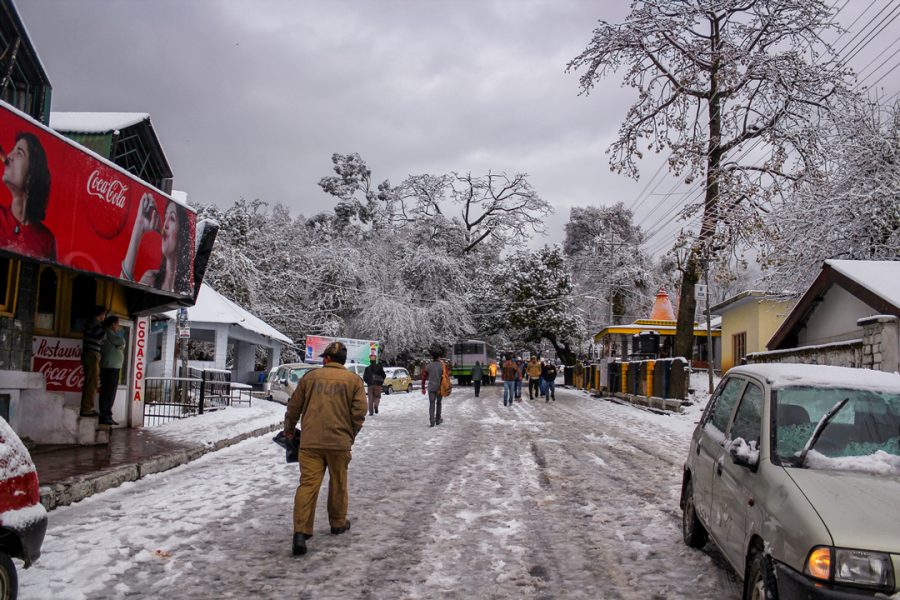 A snowy street scene in Dharamshala with pedestrians walking on the road, bare trees covered in snow, parked cars, and shop signboards including one for Coca-Cola.