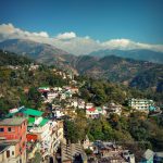 A panoramic view of Dharamshala town with multi-colored buildings nestled on green, terraced hillsides. In the backdrop, the majestic Dhauladhar mountain range rises under a blue sky with scattered clouds.