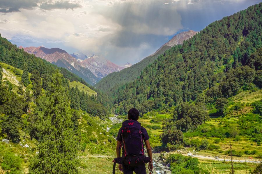 Rajgundha Valley Tour, A hiker with a backpack stands facing a lush green valley with a river flowing through it, surrounded by dense forests and towering mountains under a partly cloudy sky.