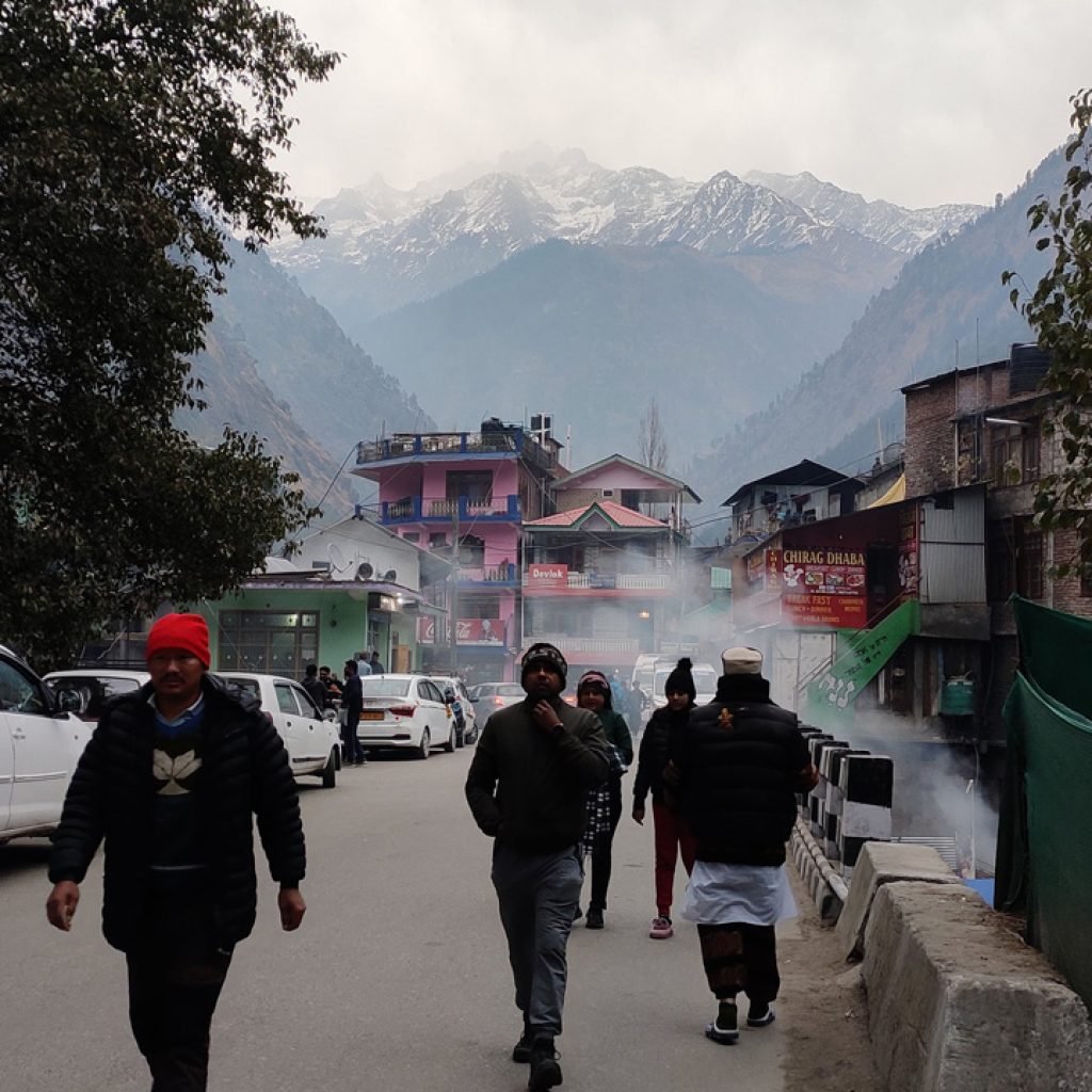 A bustling street scene in Kasol with pedestrians walking and a bus parked on the side. The backdrop features snow-capped mountains and residential buildings, with a haze that adds a mystical quality to the environment.