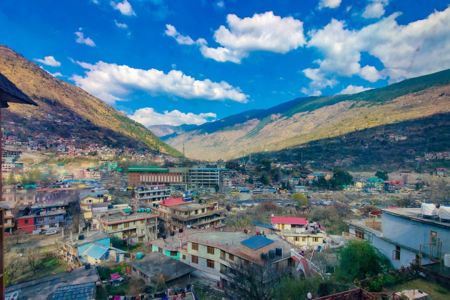 A panoramic view of Kullu Valley, showcasing a town nestled between green mountain slopes under a vibrant blue sky dotted with clouds. Buildings with colorful rooftops are spread across the valley floor, surrounded by scenic landscapes.