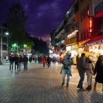 A busy night scene at Manali Mall Road with people walking along the brightly lit shopping street. The shops are illuminated with colorful lights, and visitors are seen enjoying the evening atmosphere.
