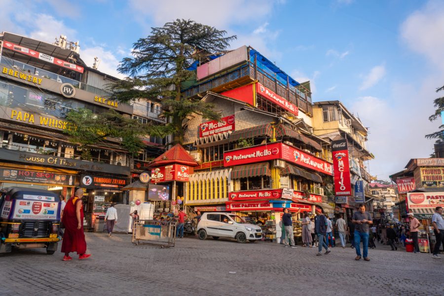 A bustling main square in McLeod Ganj with multi-story buildings featuring various shops and eateries, vibrant signage, and a mix of pedestrians and vehicles under a clear blue sky. Dharamshala Mcleodganj Package