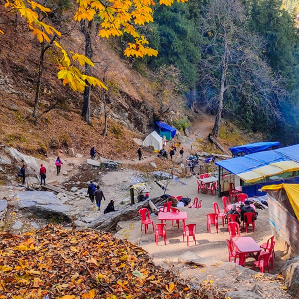 A rest stop on the Kheerganga trek with blue and yellow shelters, red chairs, surrounded by trees with autumn leaves.