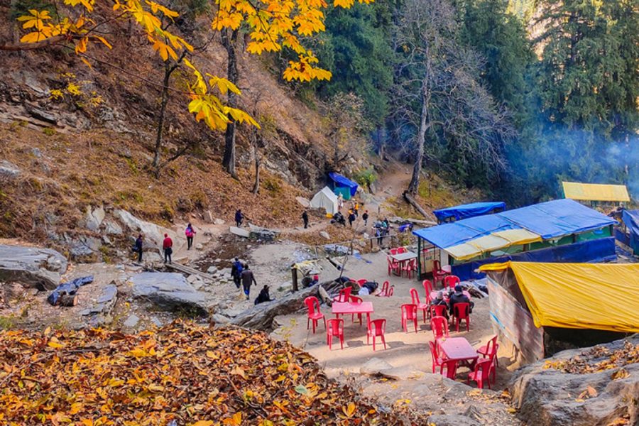 A rest stop on the Kheerganga trek with blue and yellow shelters, red chairs, surrounded by trees with autumn leaves.