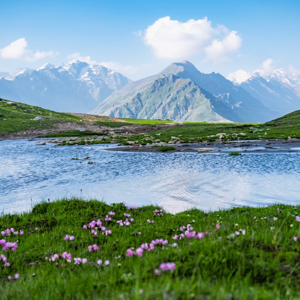 A serene view of a small lake at Rohtang Pass near Manali, with lush green grass in the foreground dotted with pink flowers, reflecting the clear blue sky. Majestic mountains rise in the background under a partly cloudy sky.