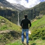 A person standing with their back to the camera, looking at the Sissu Waterfall surrounded by lush greenery and rugged mountain terrain. Manali Sissu Package