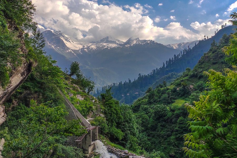 A scenic view of Tosh showcasing lush greenery, a flowing river, snow-capped mountains in the distance, and a clear sky with soft clouds. Kasol Tosh Package