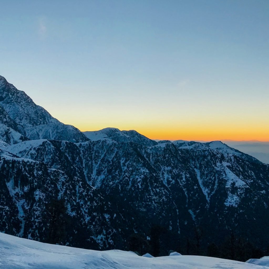 Winter sunrise at Triund top with snow-covered slopes in the foreground and the Dhauladhar mountain range in silhouette against a gradient sky.