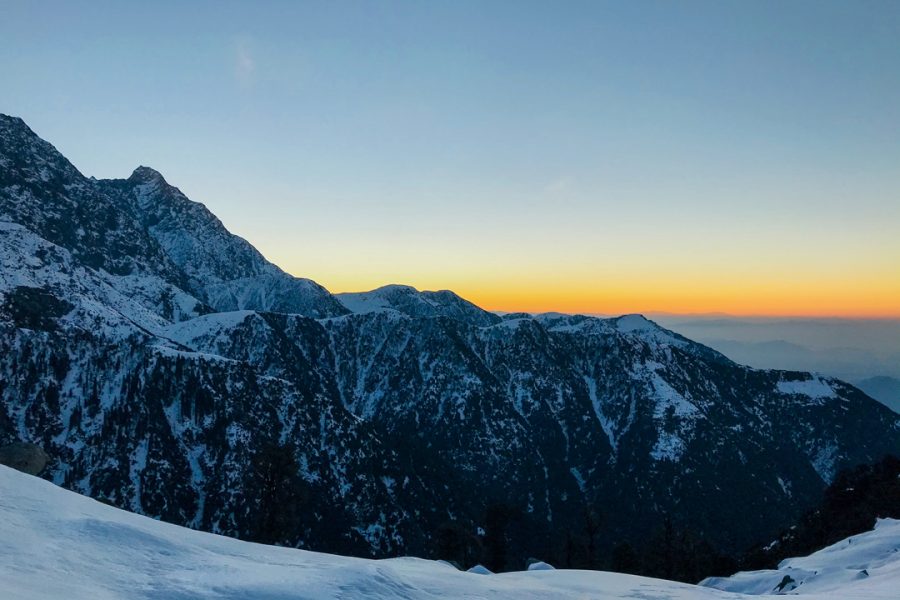 Winter sunrise at Triund top with snow-covered slopes in the foreground and the Dhauladhar mountain range in silhouette against a gradient sky.
