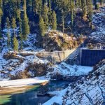 A concrete dam nestled in a snowy mountain landscape with coniferous trees and a clear blue sky.