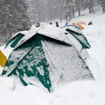 This image showcases a creatively built snowman-like figure at Juda Ka Talab base camp during the Kedarkantha trek in winter. The snow figure is adorned with a colorful scarf and a yellow hat, adding a playful touch to the scene. It stands in a snowy clearing, with snow-covered trees in the background and a blue trekking pole nearby. The surrounding tents and winter campers highlight the adventurous spirit of those exploring the remote and picturesque Kedarkantha region in the colder months.
