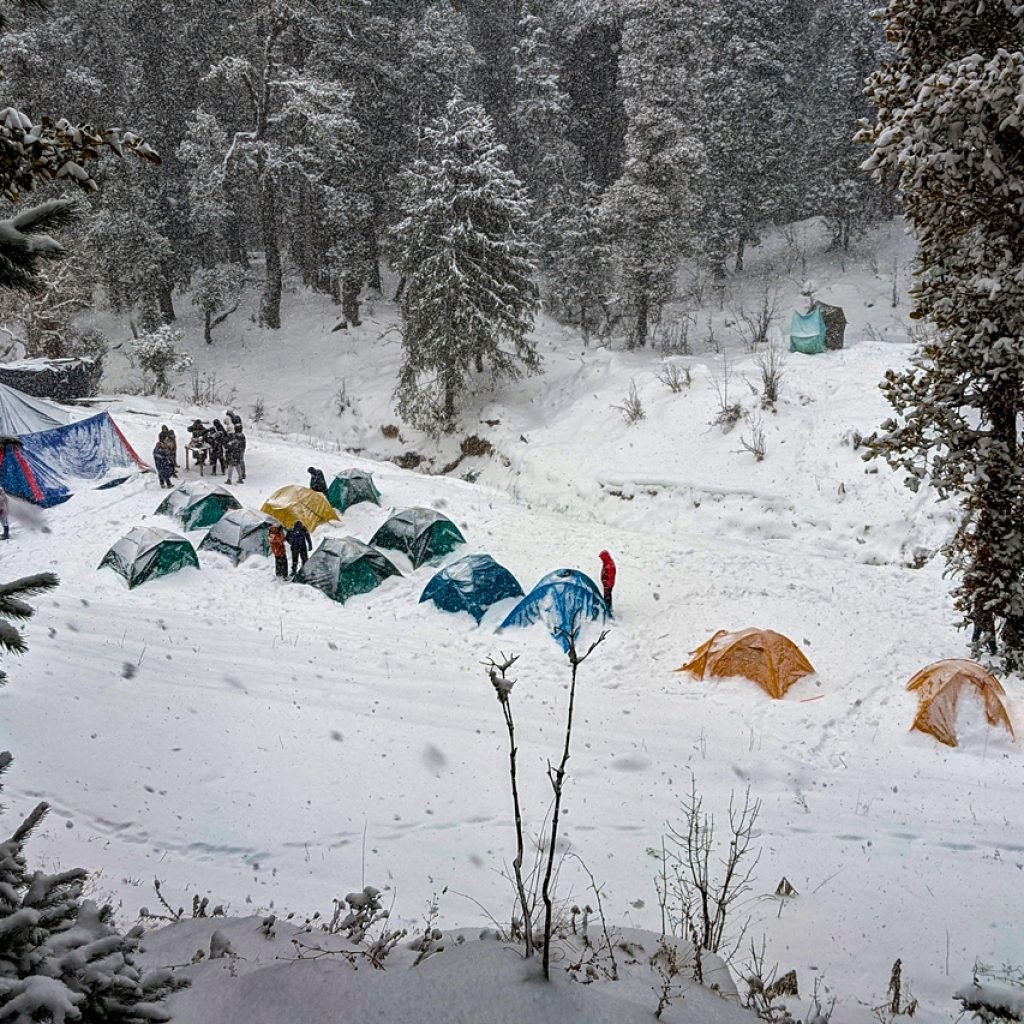 A snowy campsite at Juda Ka Talab base camp in Kedarkantha Sankri, featuring multiple colorful tents and people gathered around in the snow-covered landscape surrounded by dense forest.