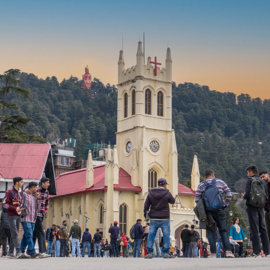 View of Christ Church on the Shimla Ridge with people walking around and a large Hanuman statue visible in the backdrop, surrounded by forested hills.