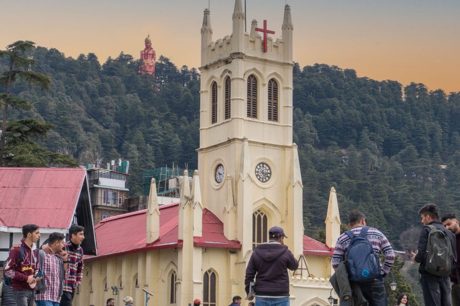 View of Christ Church on the Shimla Ridge with people walking around and a large Hanuman statue visible in the backdrop, surrounded by forested hills.