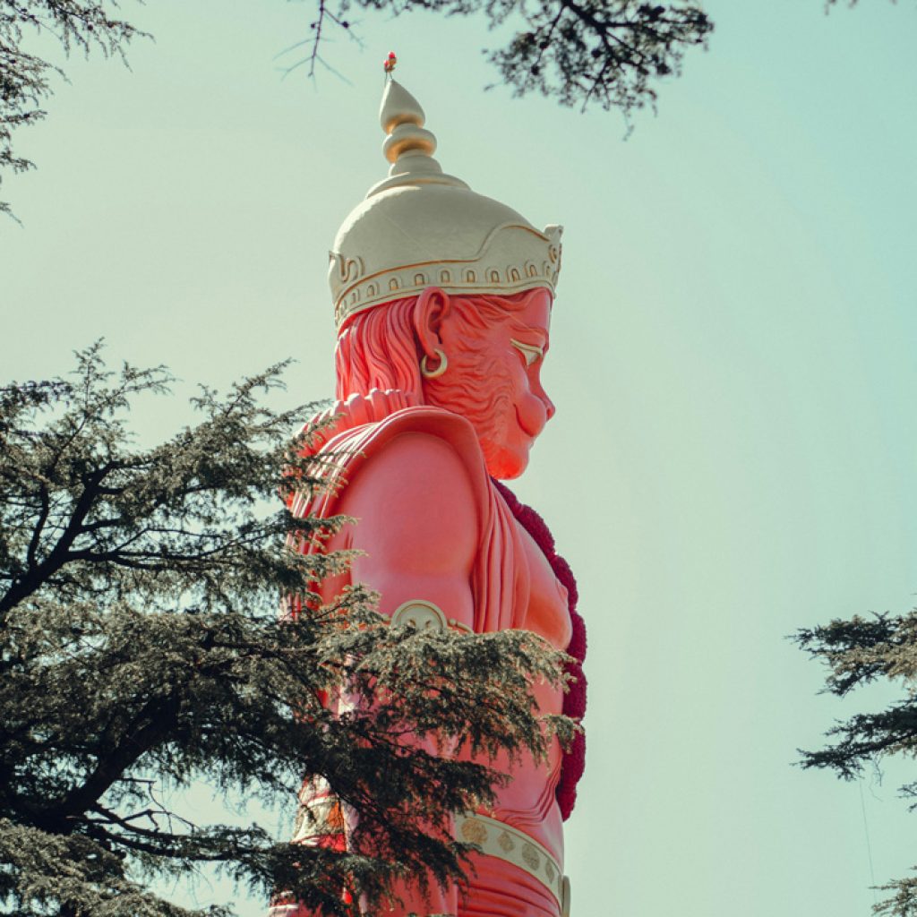 A tall red statue of Lord Hanuman, the Hindu deity, at Jakhu Temple in Shimla, partially obscured by trees under a bright sky.