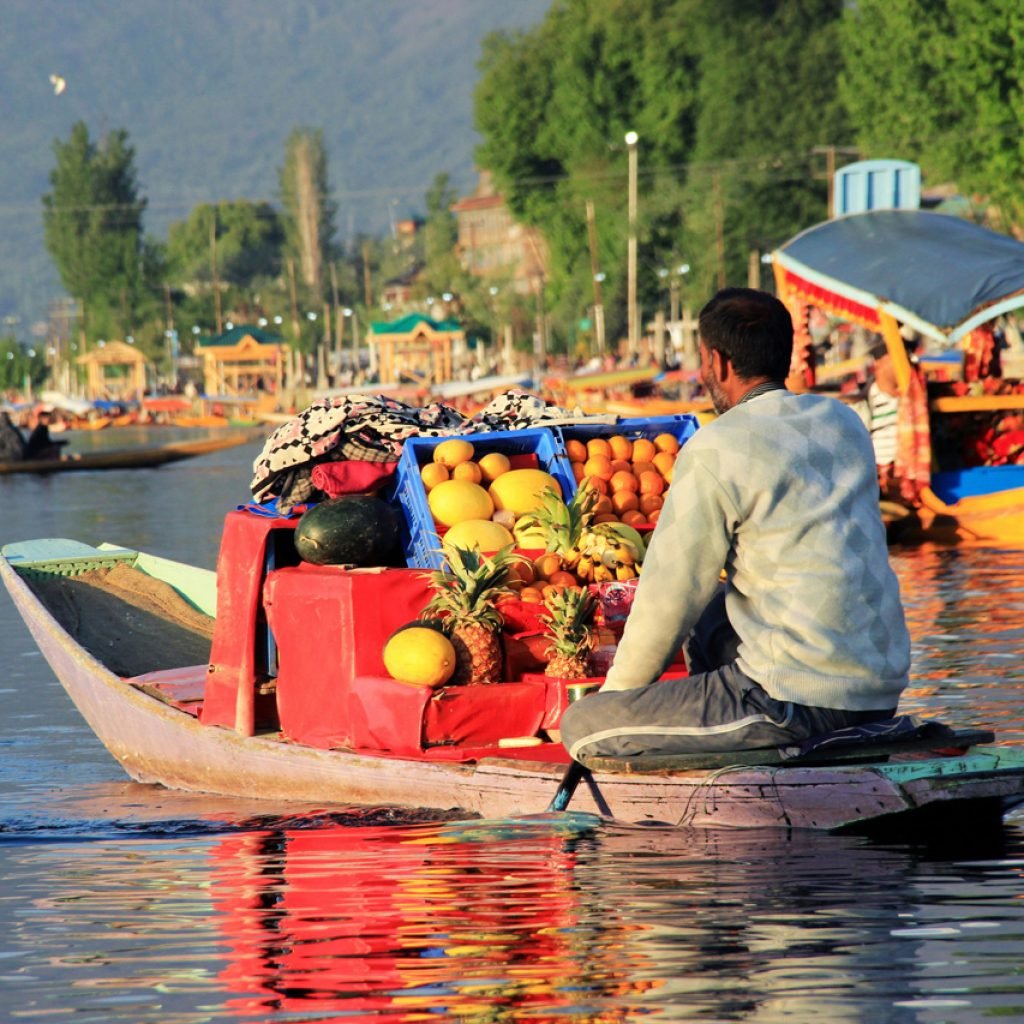 A man on a boat in a floating market on a lake, selling various fruits including pineapples, melons, and oranges. Other boats and people are visible in the background, with lush green trees and mountains surrounding the lake.