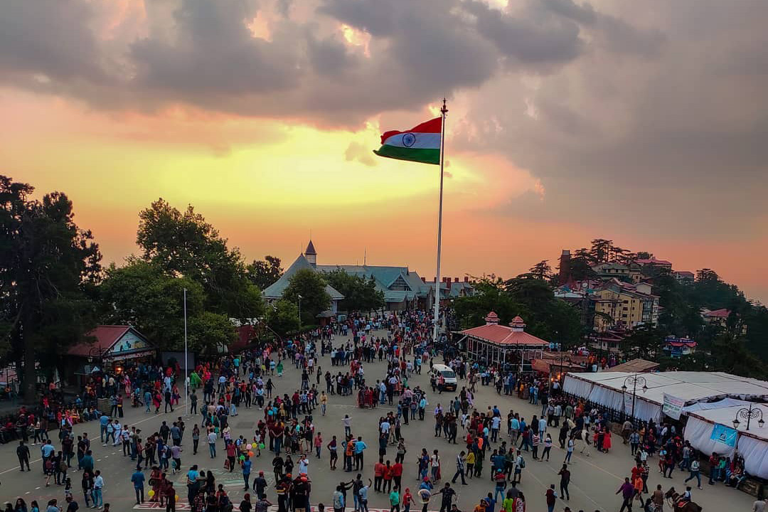 A bustling scene at Shimla Ridge with a large Indian flag waving in the foreground against a backdrop of a sunset sky. Crowds of people are gathered in the open space, surrounded by colonial-era buildings and dense foliage.