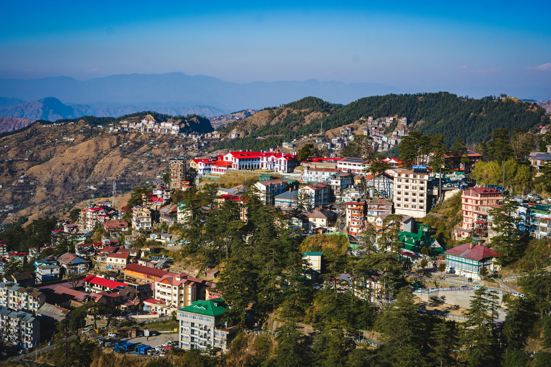 A panoramic view of Shimla town nestled in the hilly terrain with multi-storied buildings amid greenery, under a clear blue sky.